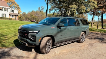 Camioneta Chevrolet Suburban verde grisácea sobre un camino de asfalto frente a una casa blanca de dos pisos, césped y árboles