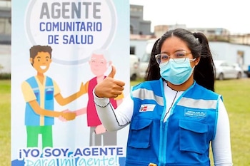Mujer peruana con mascarilla y chaleco azul de Agente Comunitario de Salud sonríe y da un pulgar arriba frente a un cartel ilustrativo, en un espacio abierto