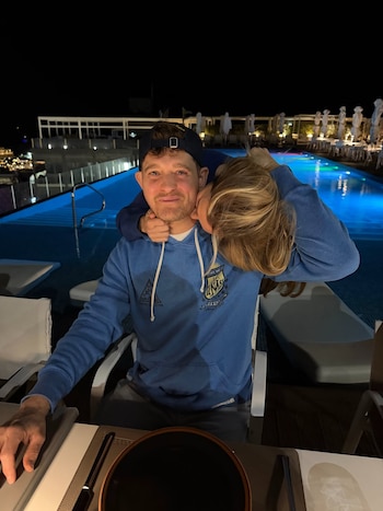 Un hombre con gorra y sudadera azul sonríe sentado a una mesa, mientras una mujer rubia lo abraza y besa su mejilla junto a una piscina
