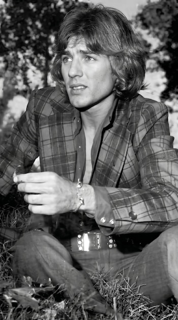Retrato en blanco y negro de un hombre joven con cabello largo, sentado en la hierba. Viste chaqueta a cuadros y camisa abierta, sosteniendo un objeto. Fondo de follaje borroso.