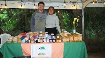 Dos personas, un hombre y una mujer, de pie detrás de una mesa con frascos de mermelada, postres, un cartel, flores y luces colgantes. Fondo verde de vegetación.