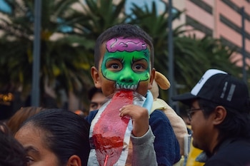 Un niño con pintura facial verde sostiene una golosina roja plana frente a su boca en medio de varias personas