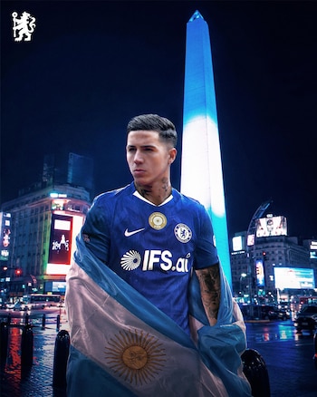 El futbolista Enzo Fernández con camiseta azul del Chelsea y bandera argentina envuelta, parado de noche frente al Obelisco de Buenos Aires iluminado en azul
