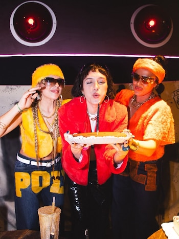 Tres mujeres con ropa colorida y piel sintética celebran un cumpleaños. La mujer del centro sopla una vela sobre una torta