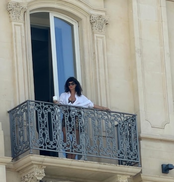 Griselda Siciliani en un balcón con baranda de metal ornamentada. Viste camisa blanca abierta, top oscuro, falda animal print, gafas de sol y sostiene una taza