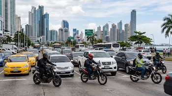 Motociclistas y automóviles transitan por una avenida de múltiples carriles con rascacielos de fondo.