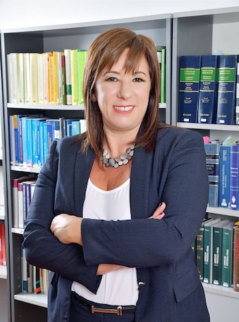 Mariana Barbitta, abogada de cabello castaño y blazer azul, sonríe con los brazos cruzados frente a un estante lleno de libros, incluyendo tomos de derecho