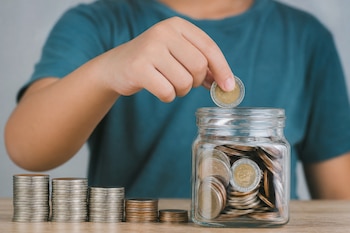 Un niño introduce una moneda en un frasco de vidrio transparente con monedas, junto a varias columnas ordenadas sobre una mesa de madera.