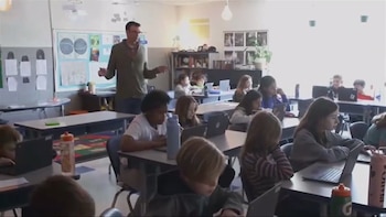 Un maestro de pie habla en un aula con varios alumnos sentados que trabajan con computadoras portátiles en sus escritorios