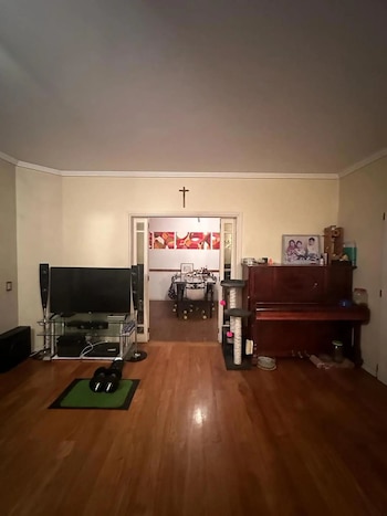 Sala de estar con piso de madera y paredes claras. Contiene un televisor, piano, rascador para gatos. Se ve un comedor por una puerta al fondo