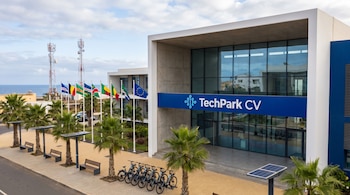 Edificio TechPark CV con fachada vidriada, palmeras, banderas de varios países, bancos con paneles solares, bicicletas eléctricas, antenas de telecomunicaciones y océano.