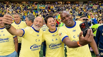 Tres personas con camisetas amarillas del Club América se toman fotografías con sus teléfonos en un estadio lleno de público.