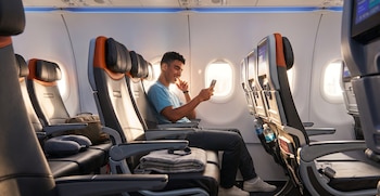 Hombre joven sentado junto a la ventanilla de un avión mirando un teléfono móvil, con asientos vacíos, almohada de viaje y manta