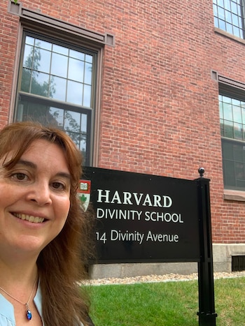 Mujer sonriente frente a un edificio de ladrillos rojos y un cartel negro con el texto "Harvard Divinity School"