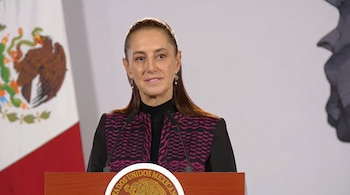 Claudia Sheinbaum frente a dos micrófonos en un atril de madera con el escudo de México y la bandera nacional al fondo.