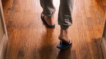 Pies y tobillos de una persona con pantalón deportivo gris y sandalias oscuras sobre un suelo de listones de madera.