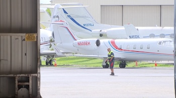 Tres aviones privados blancos con franjas azules y rojas estacionados en una pista. Un trabajador con chaleco de seguridad camina cerca. Hangar abierto con cartel de salida