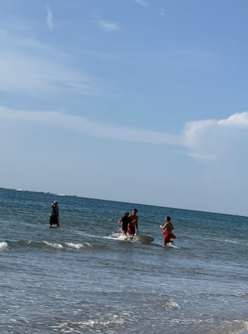 Cuatro personas en la orilla del mar, entre ellas un salvavidas con un flotador rojo y dos jóvenes junto a una tabla en el agua
