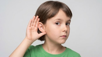 Primer plano de una niña de 8 años con cabello castaño y camiseta verde, tocándose la oreja izquierda con la mano derecha sobre un fondo blanco.