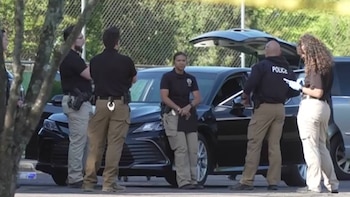 Varios agentes de policía y personal táctico están de pie junto a vehículos oscuros, uno con el maletero abierto, en un entorno exterior