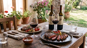 Mesa de madera con dos platos de carne asada, copas de vino tinto, cubiertos y dos botellas etiquetadas Malbec y Cabernet.
