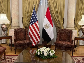 Sala diplomática: Banderas de EE. UU. y Egipto, dos sillas rojas, mesas de madera y cortinas doradas. Centro floral blanco en mesa principal