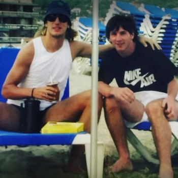 Dos hombres jóvenes están sentados en reposeras de playa; uno sostiene un mate y el otro viste una camiseta negra
