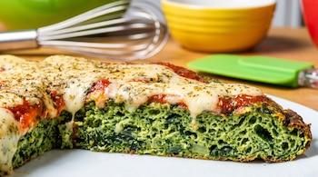 Rebanada de pizza vegetal verde con acelga, queso fundido y salsa de tomate sobre un plato blanco. Fondo con batidor y tazones de cocina.