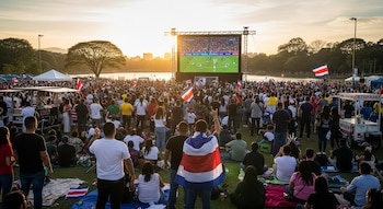 Una multitud de personas reunidas en un parque al atardecer, frente a una pantalla gigante que muestra un partido de fútbol, con banderas de Costa Rica.
