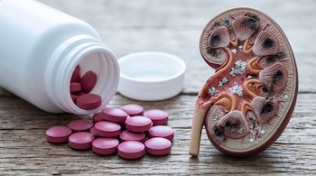 Un frasco de plástico blanco con pastillas rosas volcadas sobre madera junto a la maqueta transversal de un riñón con marcas oscuras.
