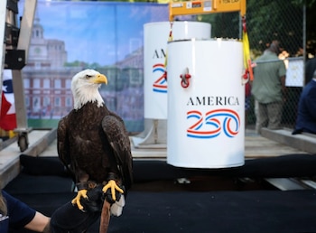 Águila calva posada en un guante. Dos cápsulas blancas cilíndricas con el logo America 250. Fondo con bandera y edificio histórico desenfocados