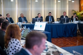 Varias personas con traje formal permanecen sentadas frente a monitores y micrófonos en una mesa de reuniones con mantel azul