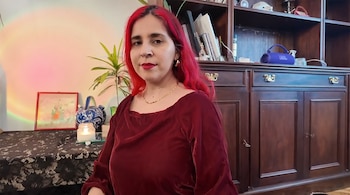 Mujer de cabello rojo con blusa granate, aretes y collar dorado, sentada frente a una pared clara con proyector de luz de arcoíris y mueble de madera oscuro
