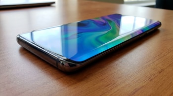 Primer plano de un smartphone plateado con una pantalla quad-curved brillante que muestra un fondo de pantalla abstracto azul, verde y púrpura.