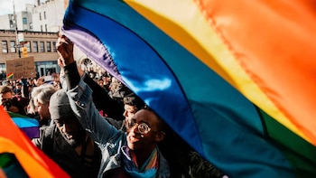 Primer plano de una multitud diversa ondeando banderas arcoíris en un evento al aire libre. Una persona con gafas redondas y un pañuelo mira hacia arriba