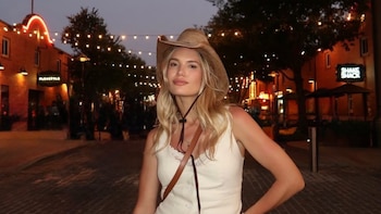 Mujer rubia con sombrero de cowboy y top blanco posa en una calle empedrada de noche, con edificios de ladrillo iluminados con luces de guirnalda de fondo