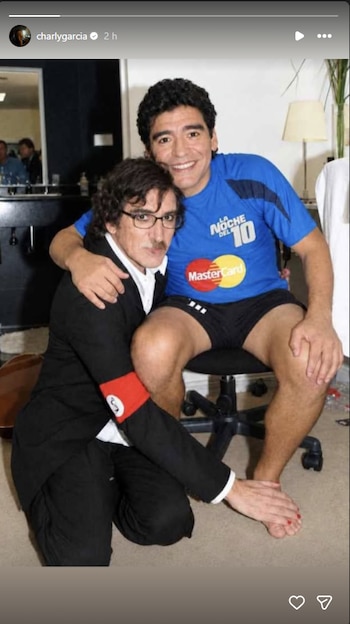 Charly García, arrodillado con traje negro, abraza a Diego Maradona, quien viste indumentaria deportiva azul y sonríe sentado en una silla