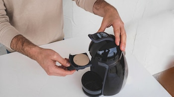 Primer plano de un hombre con suéter color crema insertando una cápsula redonda de café en el soporte de una cafetera negra sobre una superficie blanca.