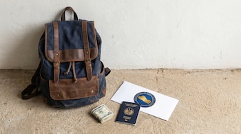 Mochila azul y marrón junto a una pared, con un fajo de dólares, un pasaporte salvadoreño y un sobre del Banco Central de Reserva de El Salvador en el suelo.