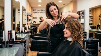 Una peluquera con tijeras y peine corta el cabello castaño de una mujer sentada frente a un espejo en un salón de belleza.