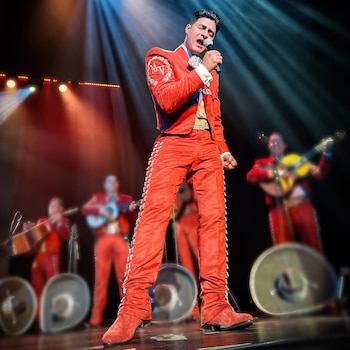 Hombre con traje de charro rojo y bordados canta con los ojos cerrados en un escenario iluminado, con otros músicos borrosos al fondo