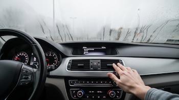 Interior de un coche con volante a la izquierda, tablero de instrumentos, rejillas de ventilación y una mano ajustando la ventilación. El parabrisas está empañado.