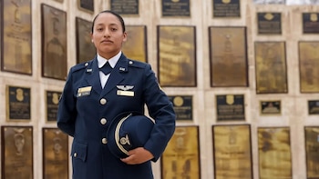 Una mujer con uniforme militar azul marino sostiene una gorra de plato frente a un muro con placas conmemorativas