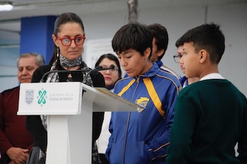 Claudia Sheinbaum Pardo con gafas rojas observa a un estudiante que lee frente a un podio con el logotipo del Gobierno de la Ciudad de México