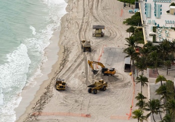 Vista aérea de tres camiones de volteo y una excavadora amarillos en una playa de arena, con el mar con olas a la izquierda, una torre de salvavidas y palmeras a la derecha