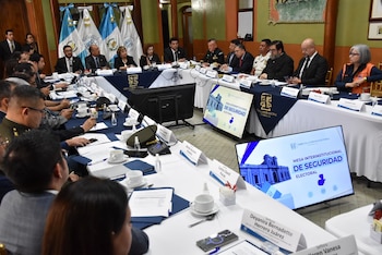 Personas en traje formal y uniformes oficiales están sentadas alrededor de una mesa en forma de herradura con micrófonos y pantallas