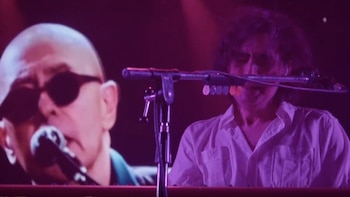 Un músico de pelo rizado toca un teclado, iluminado por luz púrpura. Detrás, en una pantalla gigante, se ve el rostro de un hombre calvo con gafas de sol