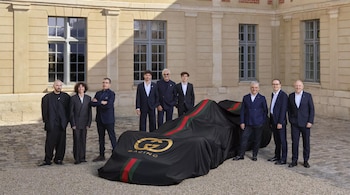 Nueve hombres y una mujer posan frente a un edificio antiguo junto a un coche de carreras cubierto con lona negra con el logo 'Gucci Racing'