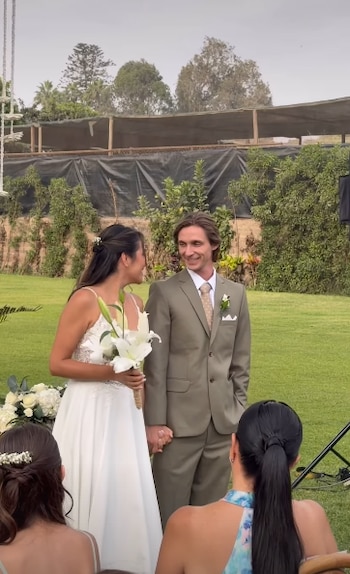 Sasha Kapsunov y Jocelyne Galarreta se miran sonriendo, tomados de la mano, en su boda al aire libre. Ella con vestido blanco, él con traje verde