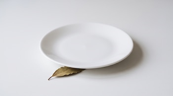 Un plato blanco redondo colocado sobre una superficie clara con una hoja de laurel seca que asoma por debajo del borde.